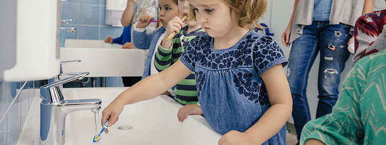 Kleinkinder putzen unter Aufsicht ihre Zähne in einem Waschraum.