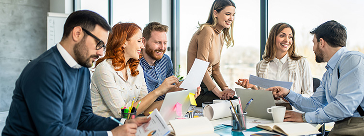 Mehrere Personen sitzen in einem Besprechungsraum an einem Tisch mit Dokumenten, offenen Laptops sowie Schreibmaterial und tauschen sich aus.