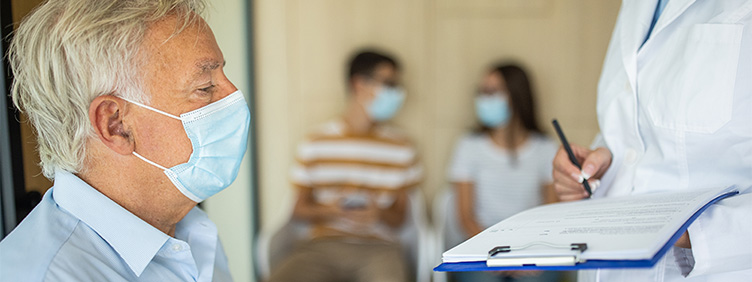 Eine Pflegeperson mit einem Formular befragt einen Herrn mit Maske, im Hintergrund warten zwei weitere Personen mit Masken.