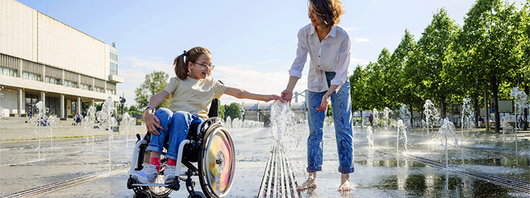 Eine Frau und ein Mädchen im Rollstuhl spielen an einem Brunnen mit Wasserfontänen in einer Stadt.