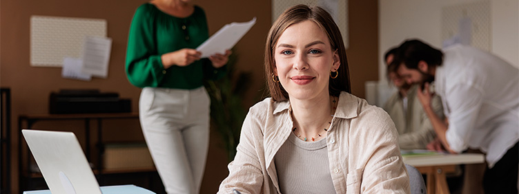 Eine Frau am offenen Laptop schaut in die Kamera. Im Hintergrund sind arbeitende Personen zu sehen.