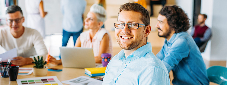 In einem Büro sitzen mehrere Personen um einen Konferenztisch mit Laptops und Schreibmaterial. Im Hintergrund befinden sich weitere Personen.