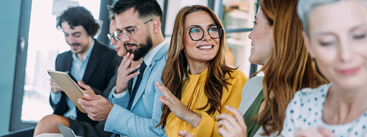 Mehrere Personen sitzen nebeneinander in einem Büroraum. Sie arbeiten auf ihrem Handy oder Laptop, andere diskutieren.