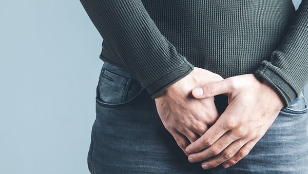 Die Körpermitte eines bekleideten Mannes, der seine Hände vor den Schritt hält.