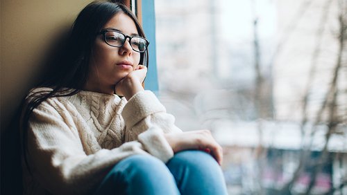 Eine Jugendliche hockt mit angezogenen Knien in der Ecke einer Zimmerwand und eines Fensters. Sie stüzt ihren Kopf auf den Arm und sieht traurig aus.