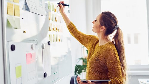Eine Frau schreibt etwas an ein Whiteboard; darauf sind neben Text auch Haftnotizen zu sehen.