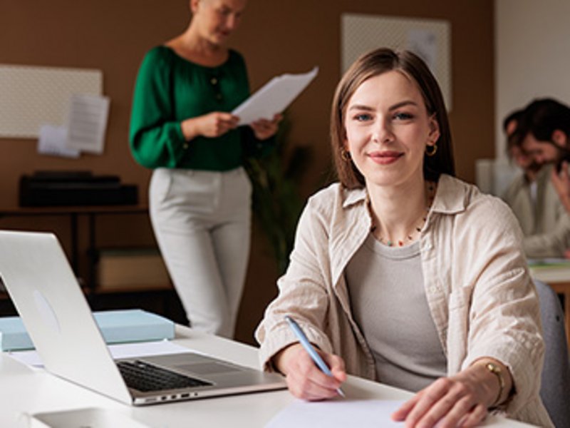 Eine Frau am offenen Laptop schaut in die Kamera. Im Hintergrund sind arbeitende Personen zu sehen.
