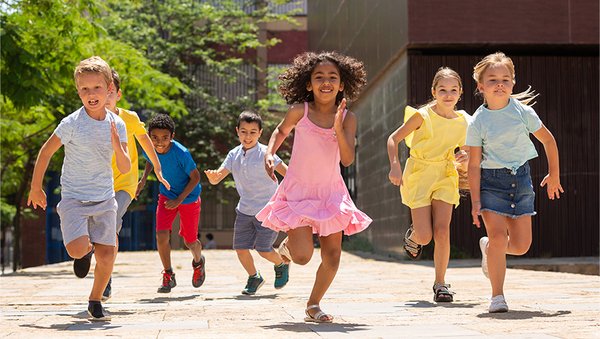 Ein paar Grundschulkinder in Sommerkleidung rennen auf die Kamera zu, im Hintergrund ein Gebäude und Bäume.