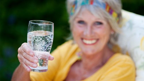 Eine Frau hält lächelnd ein Glas Wasser in die Kamera.