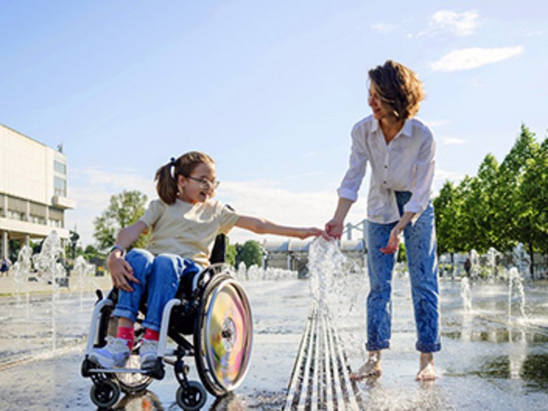 Eine Frau und ein Mädchen im Rollstuhl spielen an einem Brunnen mit Wasserfontänen in einer Stadt.