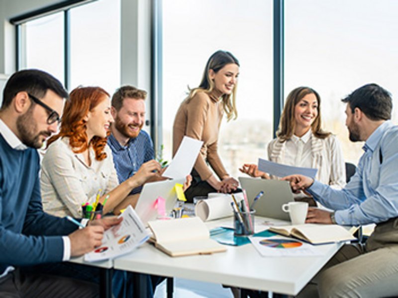 Mehrere Personen sitzen in einem Besprechungsraum an einem Tisch mit Dokumenten, offenen Laptops sowie Schreibmaterial und tauschen sich aus.