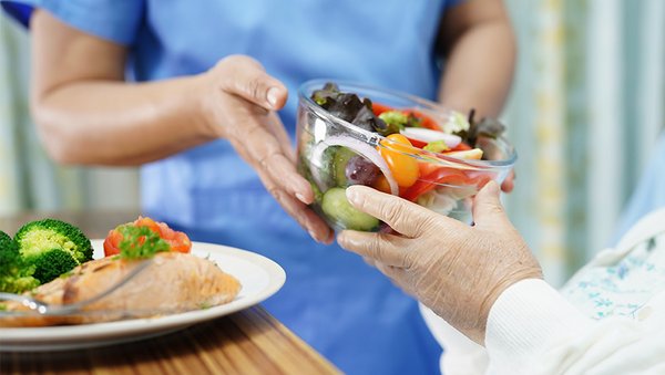 Eine Pflegekraft übergibt einer Person im Bett eine Schüssel mit Salat.