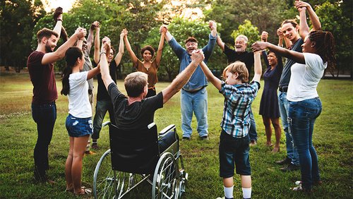 Eine Gruppe von Personen bildet auf einer Wiese in einem Park einen Kreis. Sie fassen die Hände ihrer Nachbarn und heben sie in die Luft.