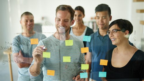 Durch eine gläserne Wand sieht man auf eine Gruppe von Personen. Sie sehen einer Person zu, die etwas auf Haftzettel schreibt, die an der Glaswand kleben.