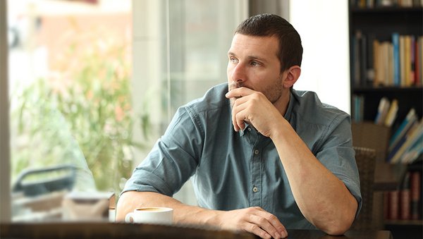 Ein Mann sitzt an einem Tisch mit einer Tasse, im Hintergrund ein Bücherregal. Er sieht nachdenklich aus dem Fenster in einen Garten.