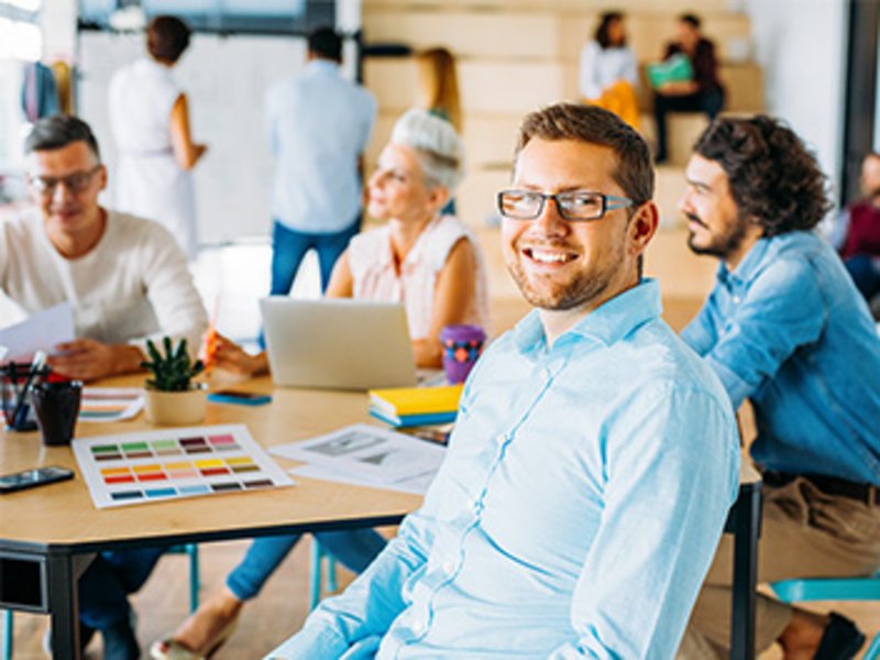 In einem Büro sitzen mehrere Personen um einen Konferenztisch mit Laptops und Schreibmaterial. Im Hintergrund befinden sich weitere Personen.