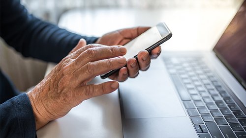 Hände einer Person am Rechner, die ein Handy halten.