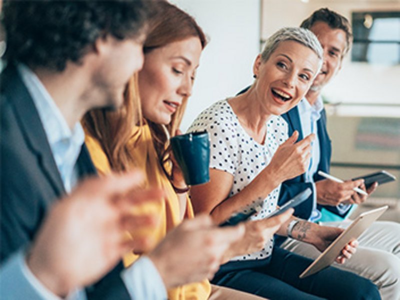 Vier Kolleg*innen sitzen nebeneinander in einem Büroraum und diskutieren lebhaft. Eine Frau hält einen Becher, ein Mann hält ein Tablet.