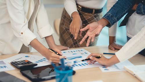 Ein Besprechungstisch mit Stiften, einem Taschenrechner und Papieren mit Diagrammen. Um den Tisch herum stehen Personen, die auf die Dokumente zeigen.