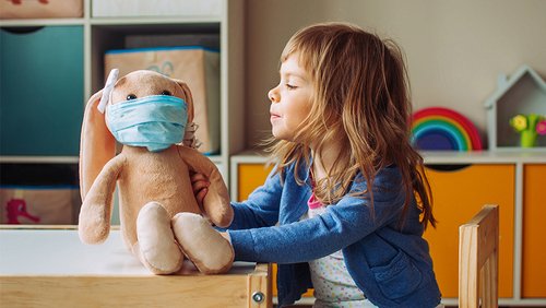Ein Kind spielt im Kindergarten mit einem Teddy, der einen Mund-Nase-Schutz trägt.