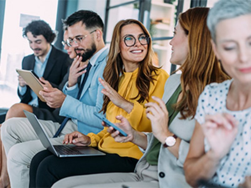 Mehrere Personen sitzen nebeneinander in einem Büroraum. Sie arbeiten auf ihrem Handy oder Laptop, andere diskutieren.