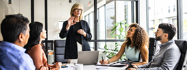 Vier Personen sitzen an einem Konferenztisch und hören dem Vortrag einer Frau zu.