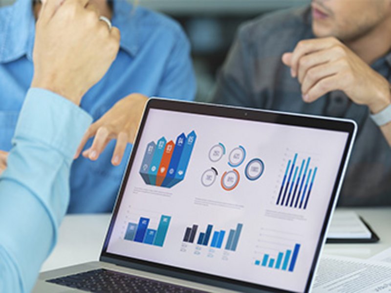 Ein Bildschirm mit Grafiken steht auf einem Tisch. Dahinter sind gestikuliernde Hände zu sehen.